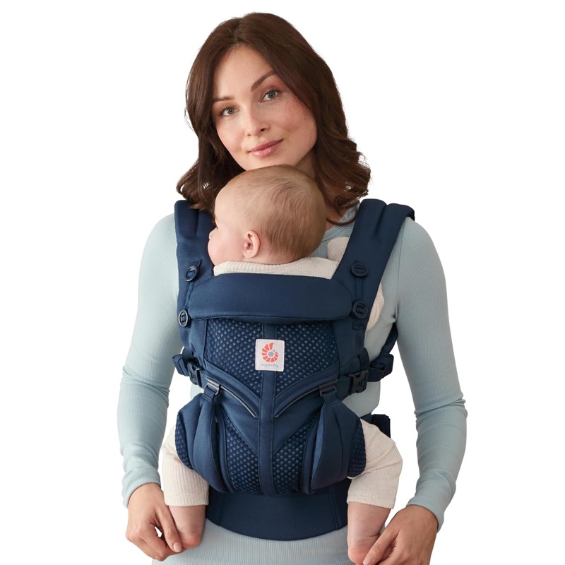 woman wearing a navy ergobaby omni carrier with a mesh panel, carrying a baby facing her chest