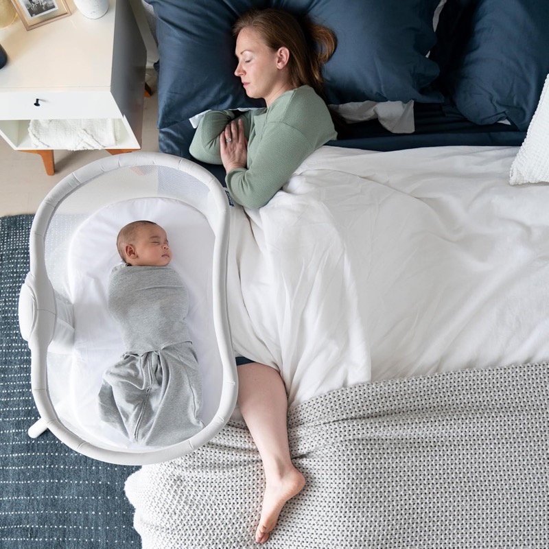 woman lying in bed next to a halo bassinest swivel sleeper with a baby asleep on its back inside