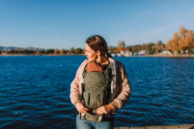 woman in a pink jacket wearing a green front carrier by a lake, with her baby's head just above the carrier panel