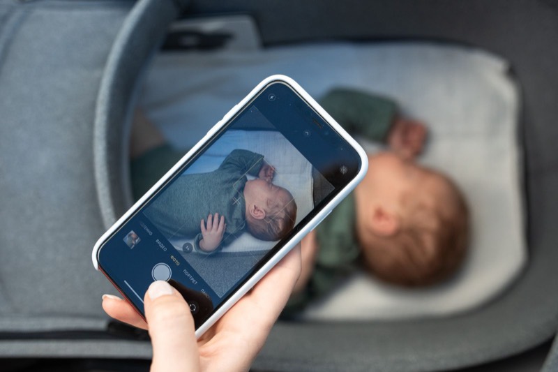 hand holding a phone that shows a live video of a baby, with the baby lying in a bassinet behind it