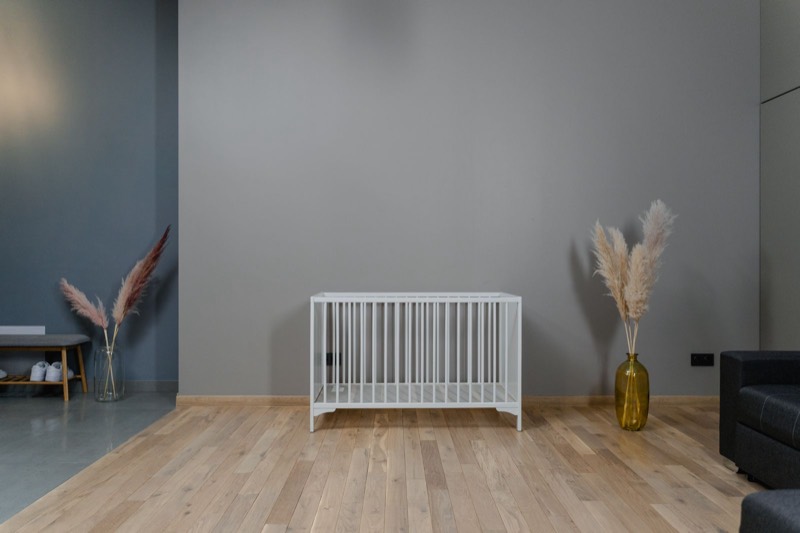 minimal nursery with a bare white crib on a light wood floor and a vase of dried grass beside it