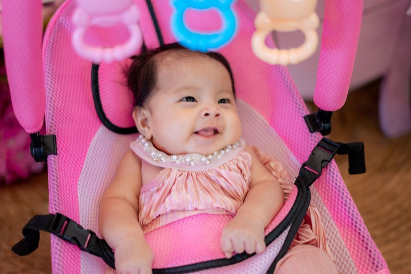 smiling baby in a pink mesh bouncer seat with the harness buckled and toys hanging above