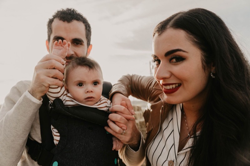 father wearing a baby in a dark front carrier with the baby's face uncovered, next to a smiling woman outdoors