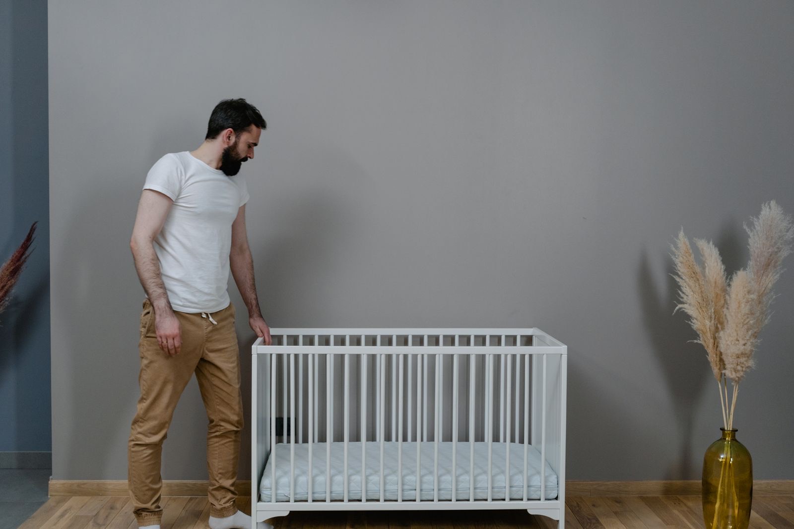man standing beside a bare white crib with a flat mattress in a minimal gray room
