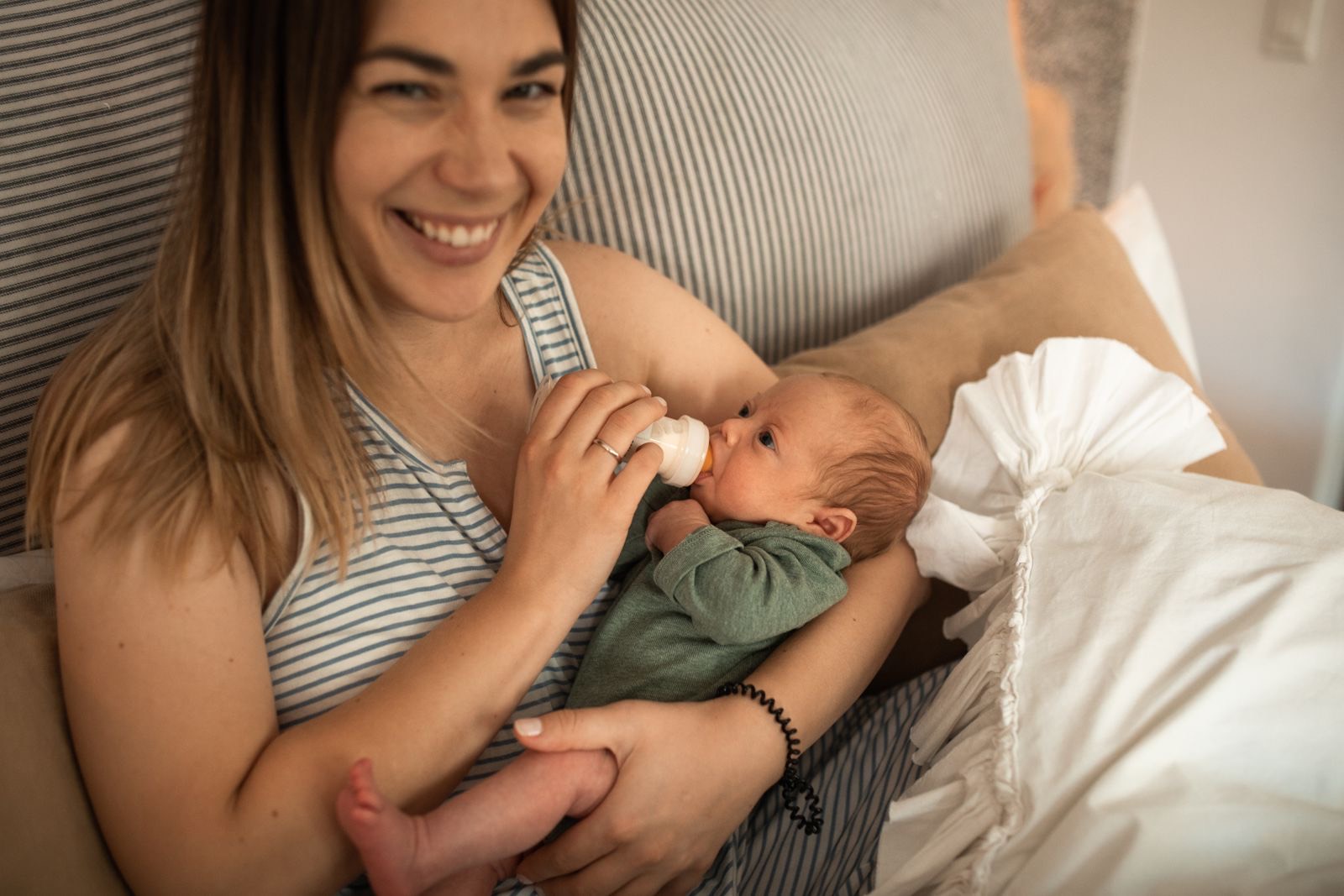 smiling mother bottle-feeding a newborn in a green outfit while sitting in bed