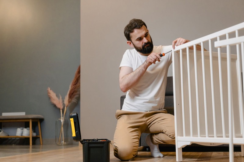 man kneeling on a wooden floor using a screwdriver to assemble a white crib, with a toolbox beside him