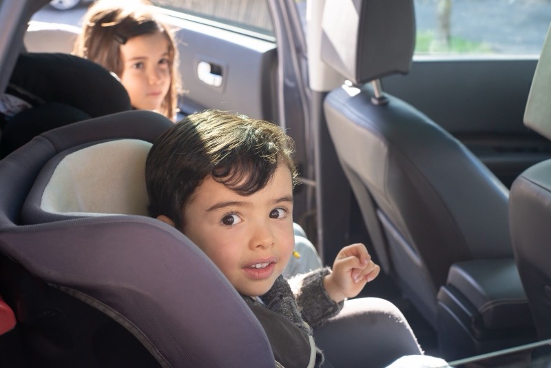 young boy sitting in a gray high-back car seat in the back of a car, with a girl in the seat behind him