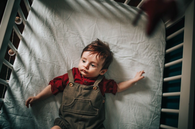 toddler lying on his back on a crib mattress with a plain fitted sheet, seen from above