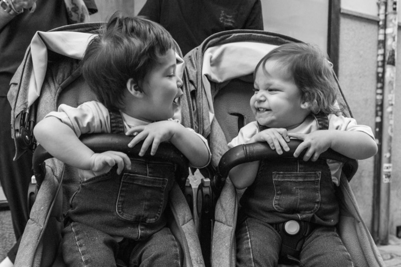 black and white photo of two toddlers sitting side by side in a double stroller and leaning toward each other