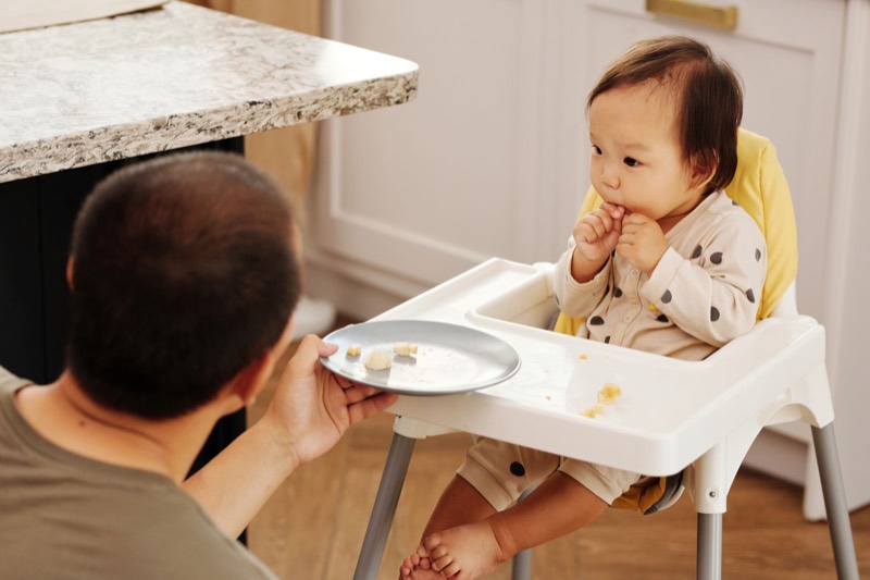 parent holding a plate and feeding a baby sitting in a white high chair with a yellow seat