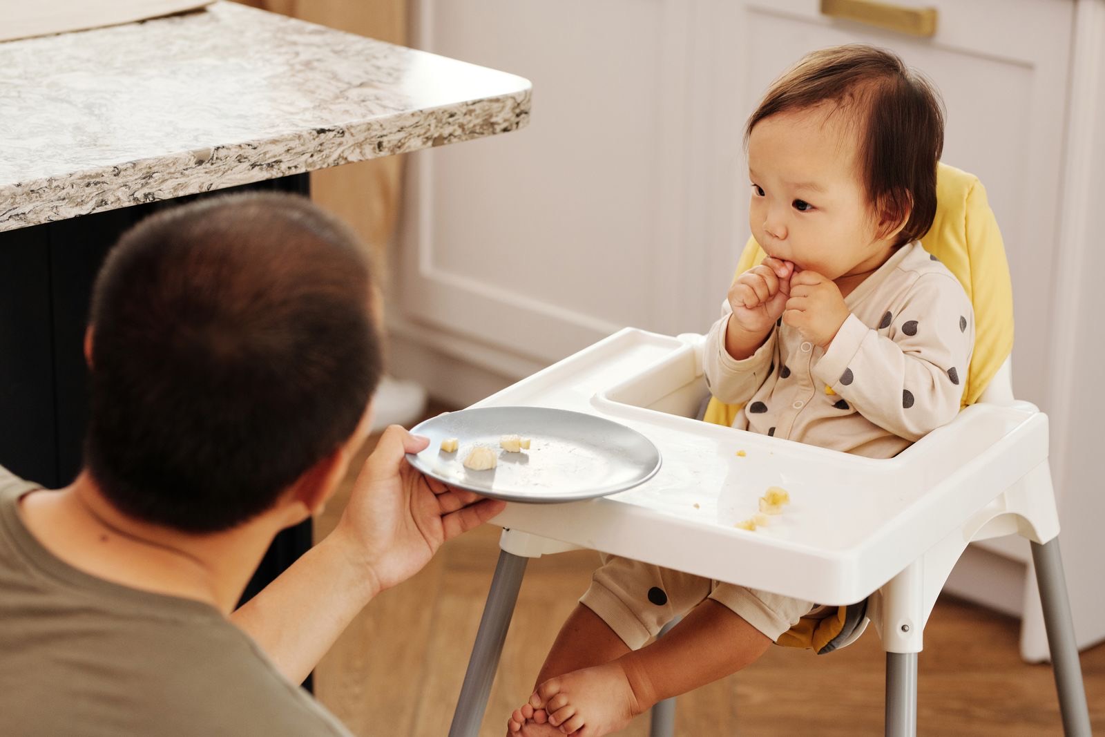 parent holding a plate and feeding a baby sitting in a white high chair with a yellow seat