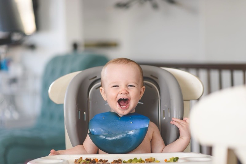 laughing baby in a gray high chair wearing a blue bib, with food on the tray