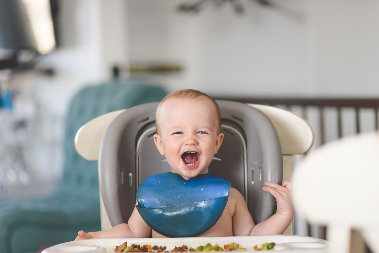 laughing baby in a gray high chair wearing a blue bib, with food on the tray