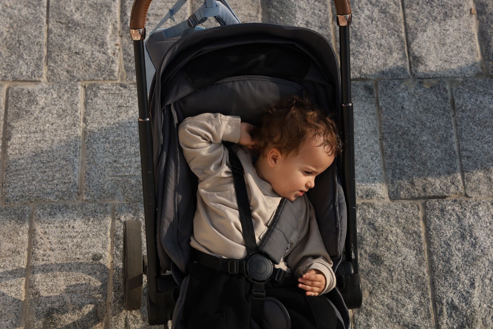 toddler in a beige sweater sitting in a black stroller with the harness fastened, seen from above on a stone pavement
