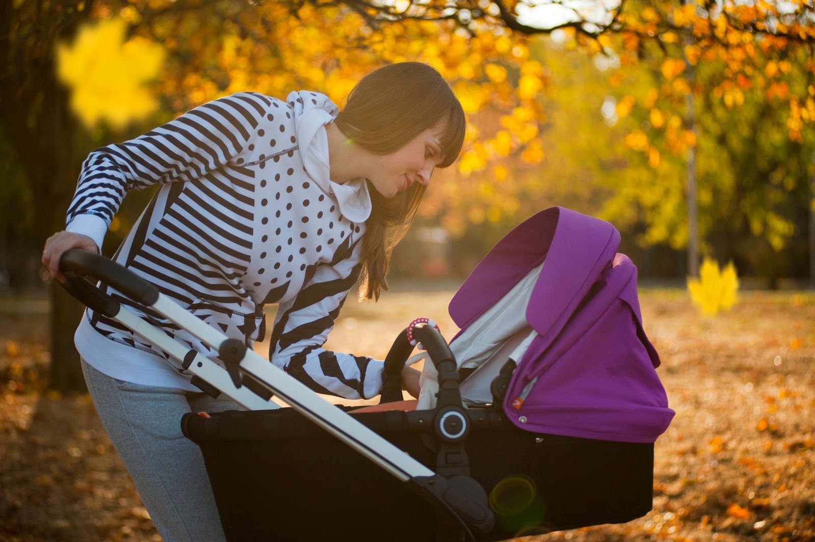 woman in a striped top leaning over a purple stroller to check on her baby in an autumn park