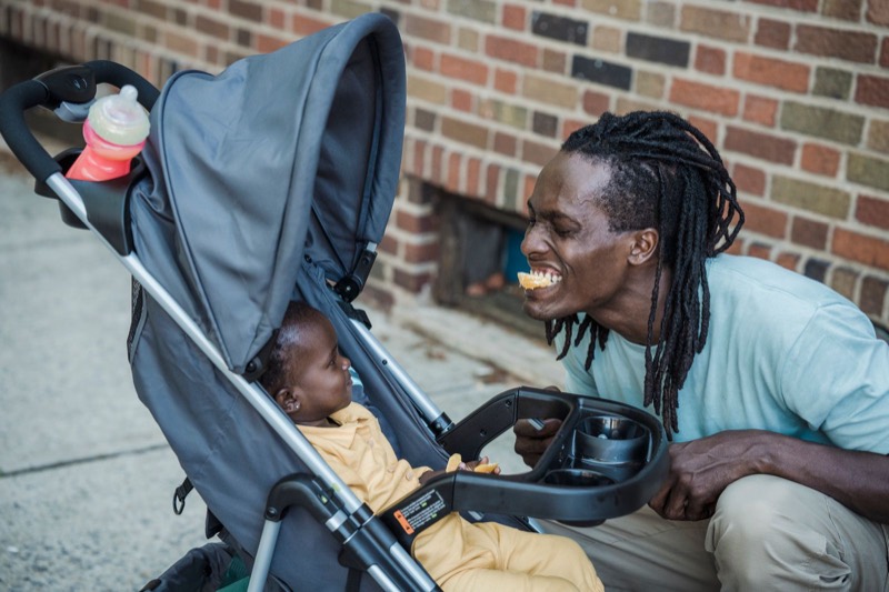 smiling father crouching beside a gray stroller to talk to his baby on a city sidewalk