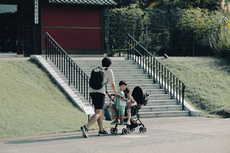 father pushing a light stroller with a child inside while another child walks beside him on a park path